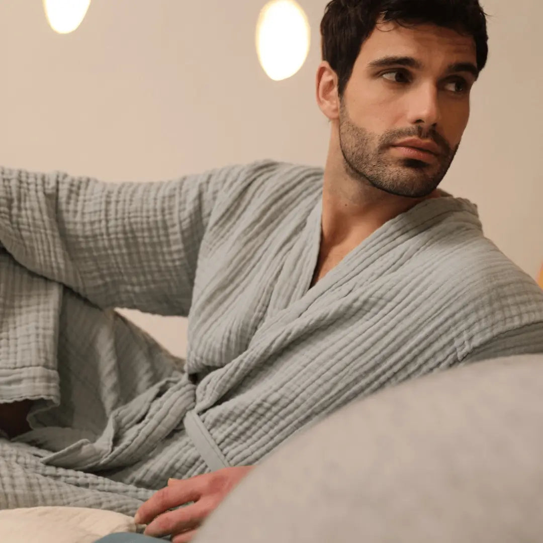 Man wearing a steel blue muslin cotton Summer Forever robe sitting on a couch in a cozy room.