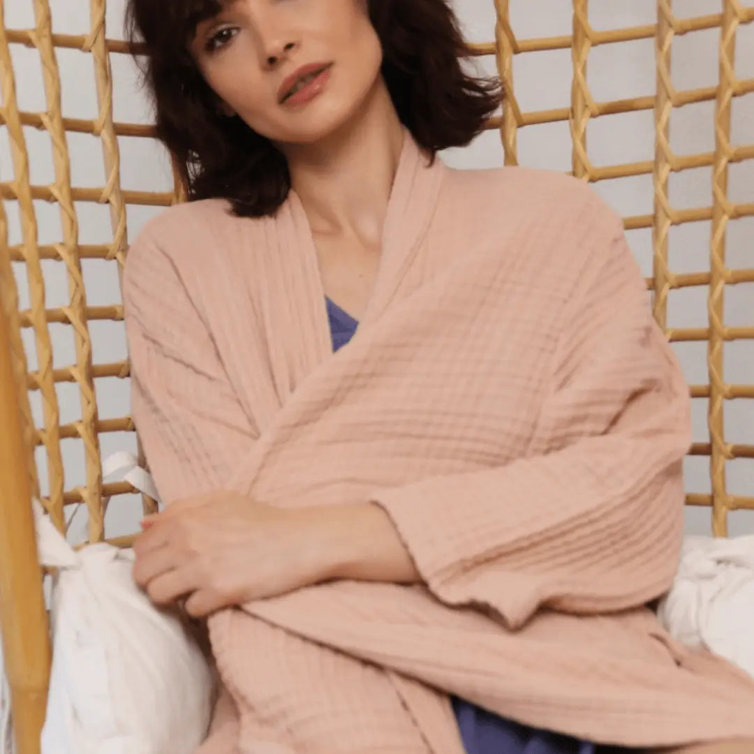 Woman wearing a pink muslin cotton  breathable Summer Forever robe relaxing nn a wicker chair.