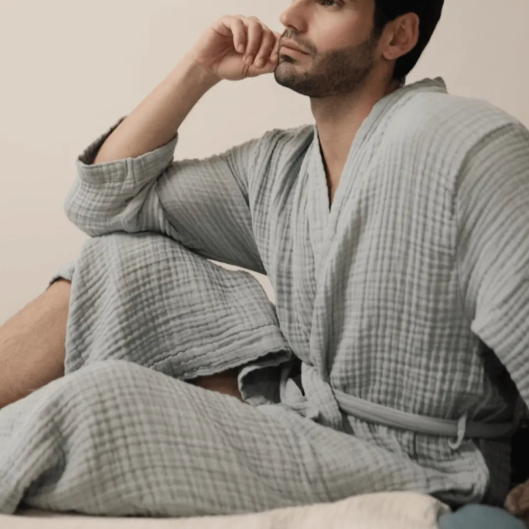 Man wearing a blue muslin cotton Summer Forever robe sitting on a bed.