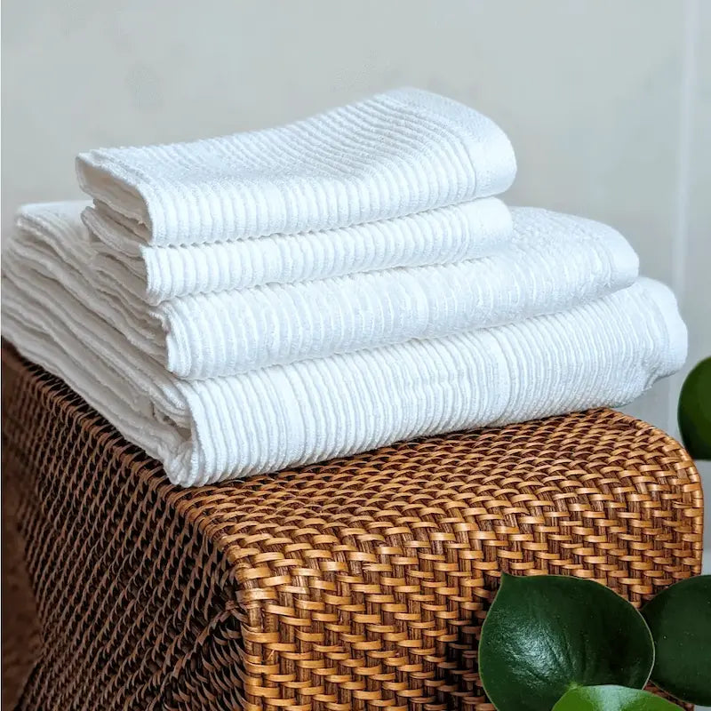 Layers of ribbed, off-white, cotton Summer Forever bath and hand towels on a wicker basket with a neutral background