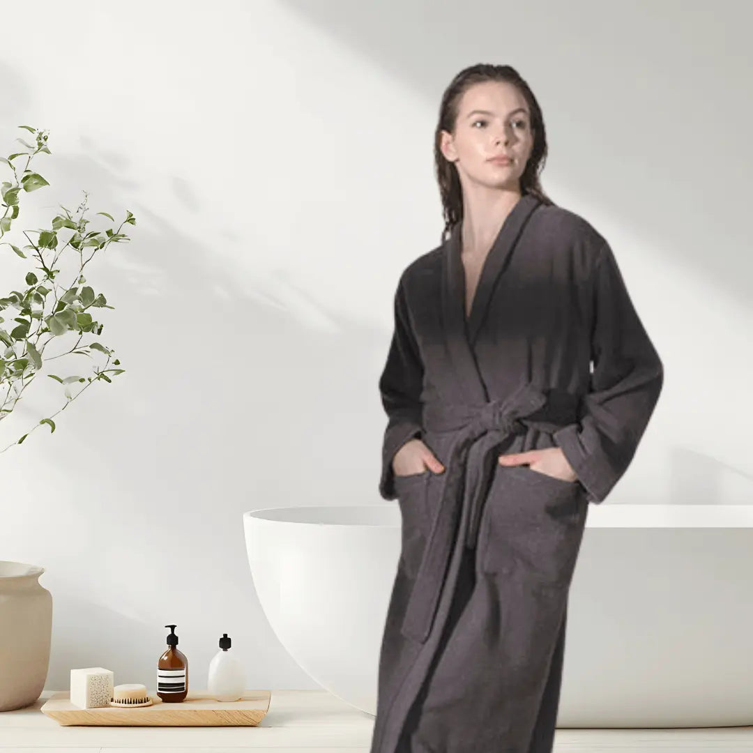Woman wearing a ribbed textured, anthracite, cotton Summer Forever robe in a bathroom setting with a plant and bathtub.