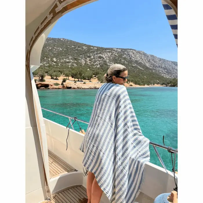 Woman on a boat wearing an extra large denim and white striped Summer Forever muslin cotton beach towel with a scenic ocean background