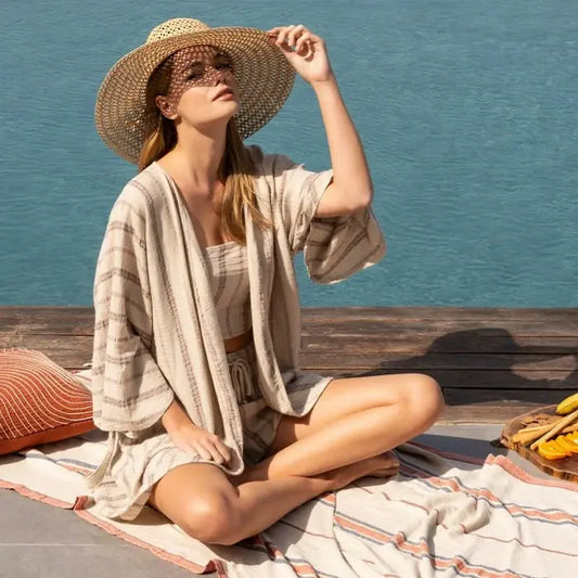 Woman in a straw hat and light dress sitting on a towel by the water.