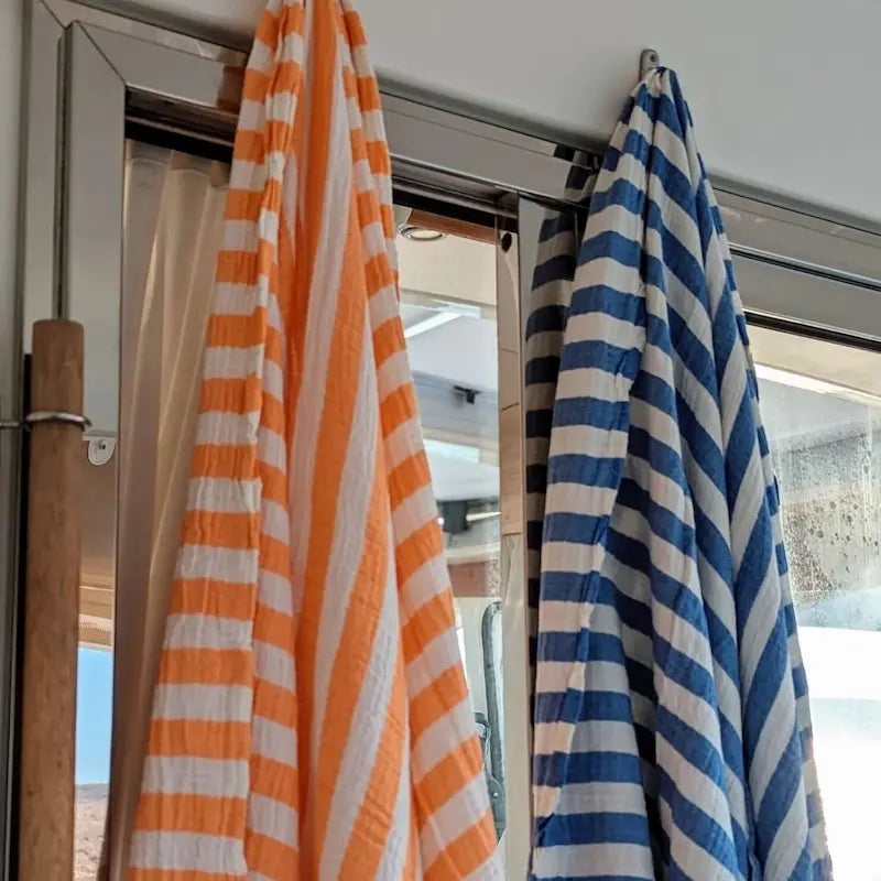 Orange and white striped Summer Forever beach towel, blue and white striped muslin beach towel hanging on a boat rack with a window in the background.