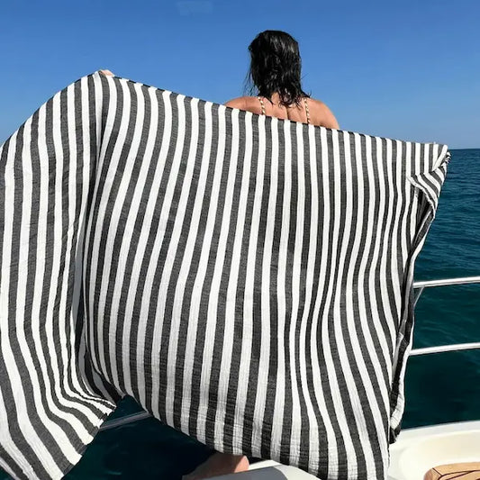 Person sitting on a boat with an extra large Summer Forever black and white striped cotton beach towel draped over them, looking out at the ocean.