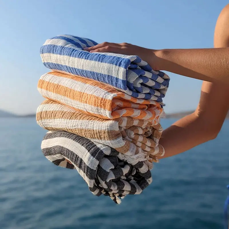 Stack of colorful striped Summer Forever Muslin cotton extra large beach towels held by a person with a blurred blue lake background